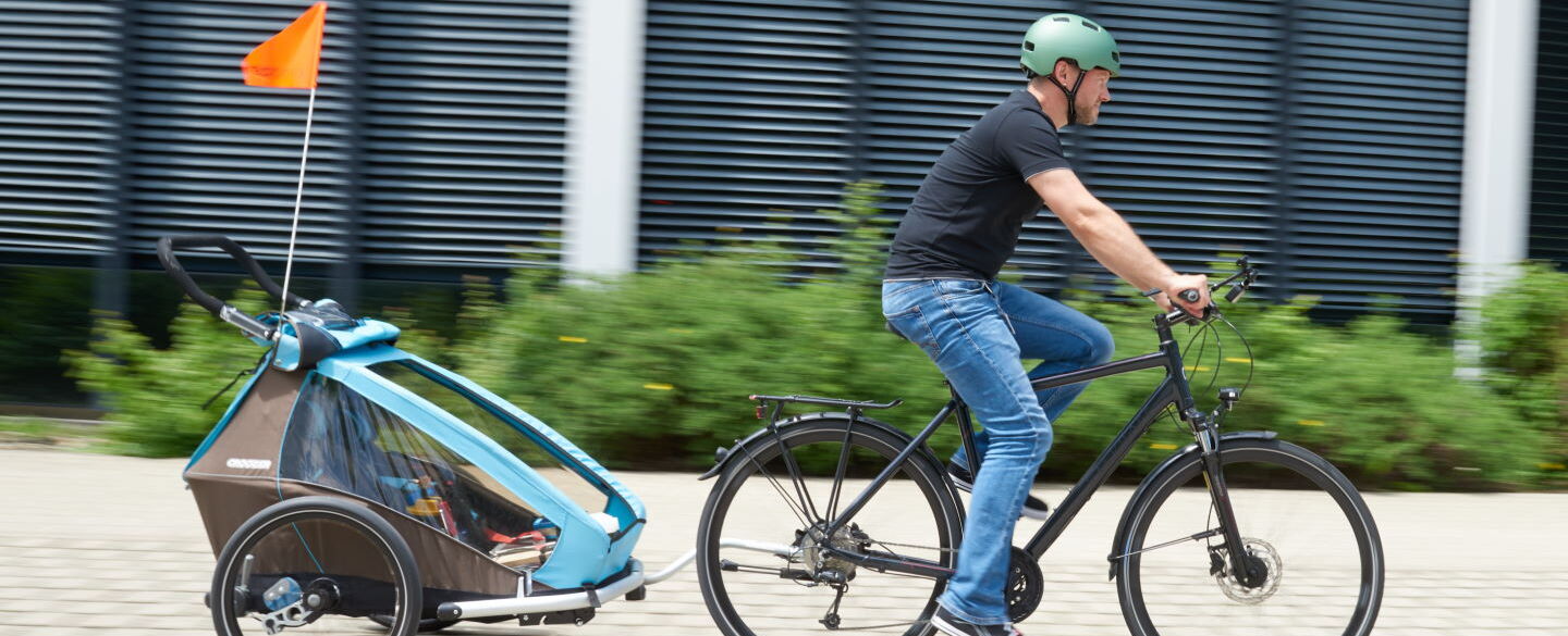 FahrradKiTra 2021 Ein Mann fährt mit einem Fahrrad auf einem gepflasterten Weg. Er trägt einen türkisen Helm, ein schwarzes T-Shirt und blaue Jeans. An seinem Fahrrad ist ein Kinderfahrradanhänger befestigt, in dem sich ein Kind befindet.
