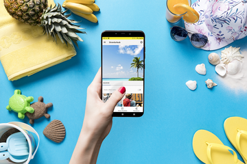 Meine Reise App - Strandurlaub Eine Hand hält ein Smartphone mit einem Strandurlaubbild, umgeben von Sommerutensilien: Handtuch, Ananas, Sonnenhut, Sonnenbrille, Sandspielzeug, Muscheln und Flip-Flops.