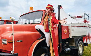 Ein Mann in einem Rot-weißen Kostüm eines Zirkusdirektors steht am Kotflügel eines alten, roten MAN-Zugfahrzeugs. In Hintergrund das Zirkuszelt.