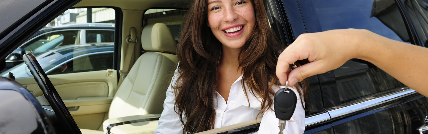 Autokauf Junge Dame sitzt in neuen Auto und bekommt Autoschlüssel übergeben.