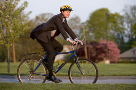 Fahrrad Radfahren in die Arbeit Businessman Ein Mann im schwarzen Anzug mit umgehängter Aktentasche und gelbem Fahrradhelm fährt auf einem Fahrrad.